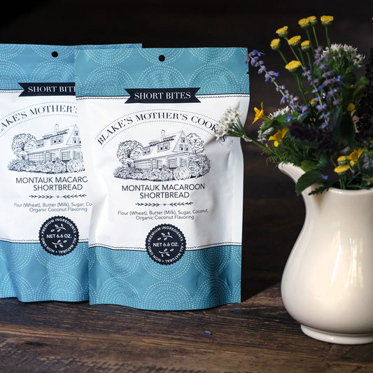 Two 6.6 oz. Blake's Mother's Cookies Shortbites Pouches sitting on a table with country flowers in an ivory vase