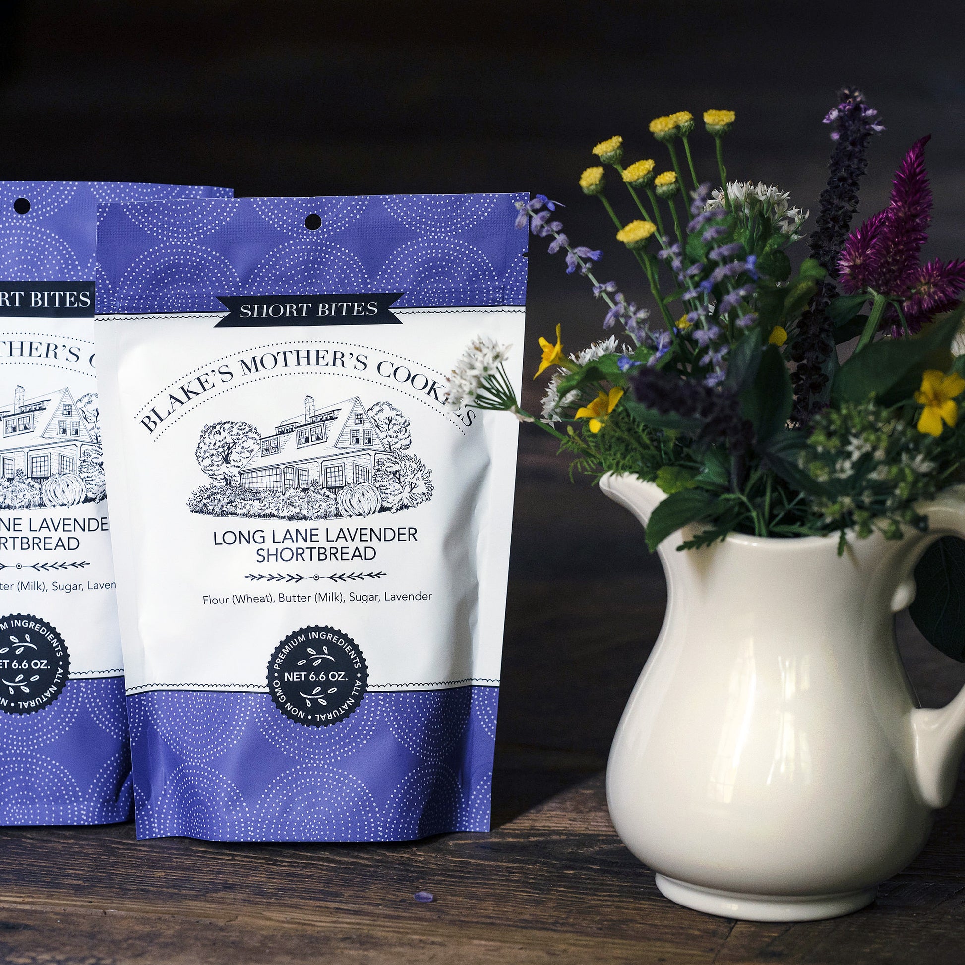 Two pouches of Blake's Mother's Cookies Long Lane Lavender Shortbites with a creamy vase filled with country flowers sitting on dark wooden table.