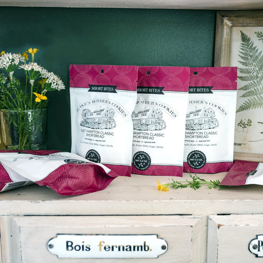 Six 2.2 oz. pouches of Blake's Mother's Cookies Short Bites sitting on a painted wooden shelf with country flowers