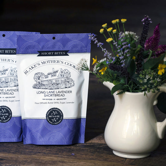 Two pouches of Blake's Mother's Cookies Long Lane Lavender Shortbites with a creamy vase filled with country flowers sitting on dark wooden table.