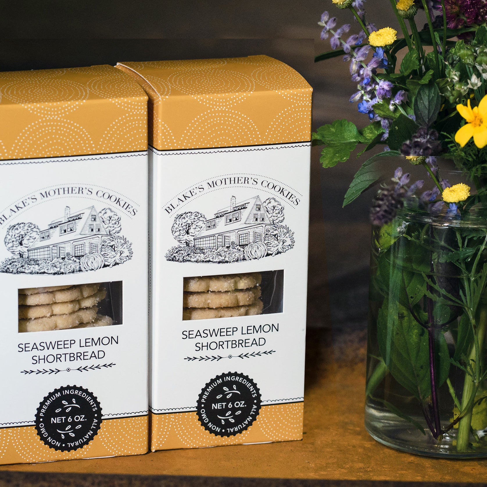 Two colorful golden yellow boxes of Sea Sweep Lemon Shortbread sitting on a wood table with a vase of blue and yellow country flowers
