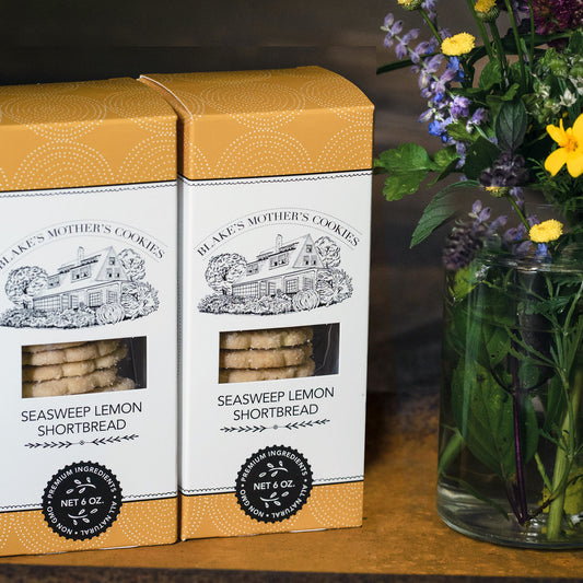 Two colorful golden yellow boxes of Sea Sweep Lemon Shortbread sitting on a wood table with a vase of blue and yellow country flowers