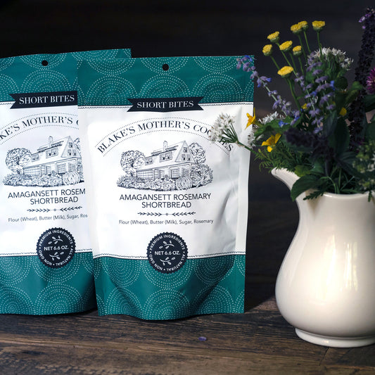 Two large 6.6 oz. pouches of Blake's Mother's Amagansett Rosemary Cookies sitting on a table with a pitcher of country flowers next to the pouches.