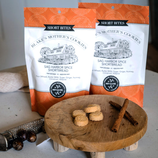 Two pouches of Blake's Mother's Cookies Sag Harbor Spice Short Bites with a Bites on a wooden serving platter and surrounded by cinnamon and nutmeg.